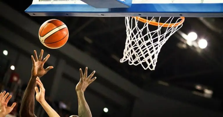 photo of the back of a basketball backboard with several athletes hands reaching up for a basketball as it heads towards the hoop