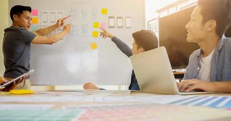 photo of 3 men working on a project together in front of a white board that is covered in doodles and post-it notes to symbolize digital product development