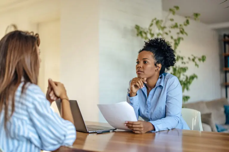 Two women having a serious conversation across a table, with one woman holding a paper and the other listening attentively. A laptop is open on the table all to represent how to answer why do you want to leave your current job in an interview.