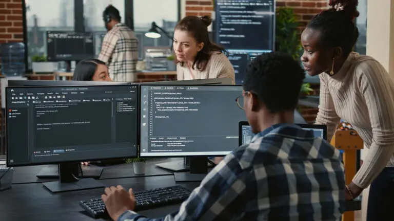 A team of diverse software developers collaborates in a modern office. One developer is seated at a dual-monitor setup, coding on both screens. Three colleagues stand nearby, engaged in a discussion while reviewing the code on the monitors. The workspace features large windows, brick walls, and several computer stations, creating an open and tech-focused environment.