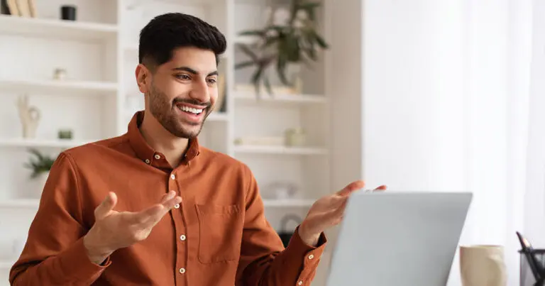 A young man in a rust-colored shirt is engaged in a virtual interview, smiling and gesturing while looking at his laptop. He is seated in a bright, well-lit room with shelves and plants in the background, creating a professional yet comfortable environment.