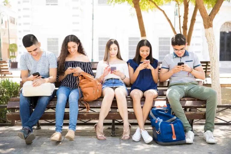 Five generation alpha boys and girls sitting on a park bench happily looking at their phones together.