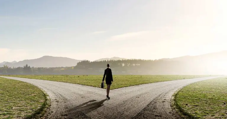 A woman in business attire stands at a fork in a dirt road, contemplating which path to take. She holds a briefcase in her left hand and faces the sunrise in the distance, with misty mountains and a line of trees visible on the horizon. The scene symbolizes decision-making and choosing a direction in life or career.