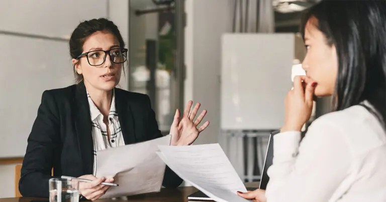 The image shows a businesswoman in a professional setting, engaged in a serious conversation with another woman. The businesswoman, wearing glasses and a dark blazer, is holding a piece of paper and gesturing with her hand, emphasizing a point. The other woman, who has long dark hair, is sitting across the table, attentively listening with a thoughtful expression. The background includes a whiteboard and a laptop, indicating an office or meeting room environment.