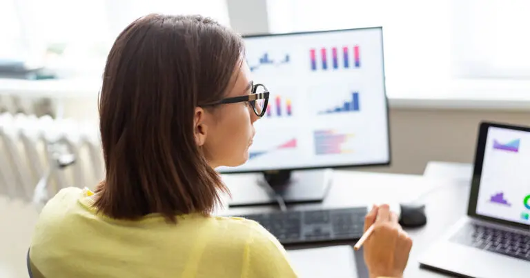 A woman wearing glasses is sitting at a desk, analyzing data displayed on a large monitor and a laptop. The screens show various colorful bar graphs, line charts, and pie charts, indicating she is working with predictive analytics. The setting appears to be a bright, modern office.