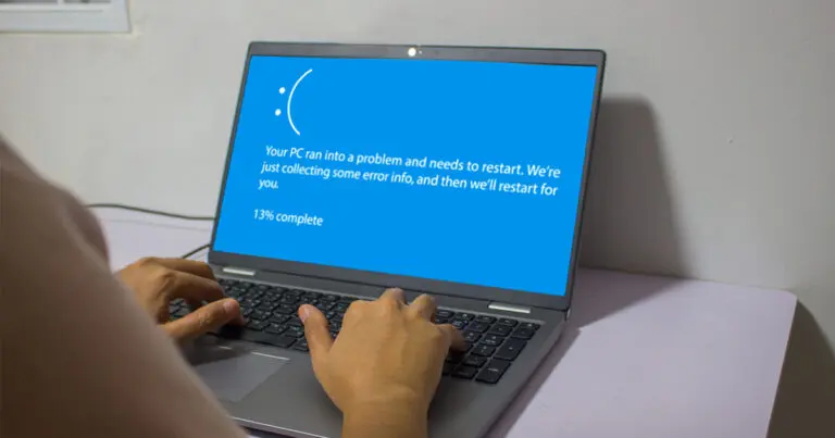 A laptop screen displaying a blue error screen with a sad face icon and the message 'Your PC ran into a problem and needs to restart. We're just collecting some error info, and then we'll restart for you.' The progress is shown as 13% complete. A person's hands are visible on the keyboard.