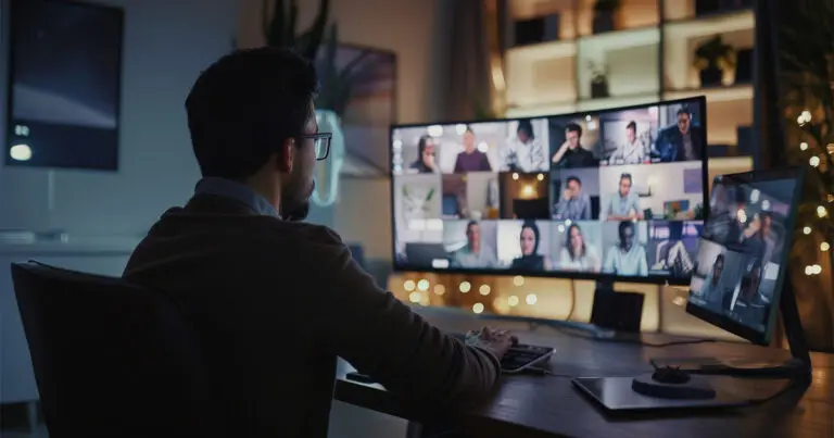 A man sits at his desk in a dimly lit room, engaging in a virtual meeting. He faces multiple computer monitors displaying a grid of video call participants, each appearing in their own separate window. The background features shelves with various decorations and soft, ambient lighting, creating a warm and focused workspace environment.