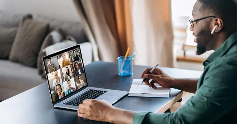 A man wearing glasses and earbuds is sitting at a desk, participating in a virtual meeting on his laptop. The laptop screen shows a video call with multiple participants. He is taking notes on a notepad with a pen, and the background features a cozy, well-lit living room with a sofa and curtains.