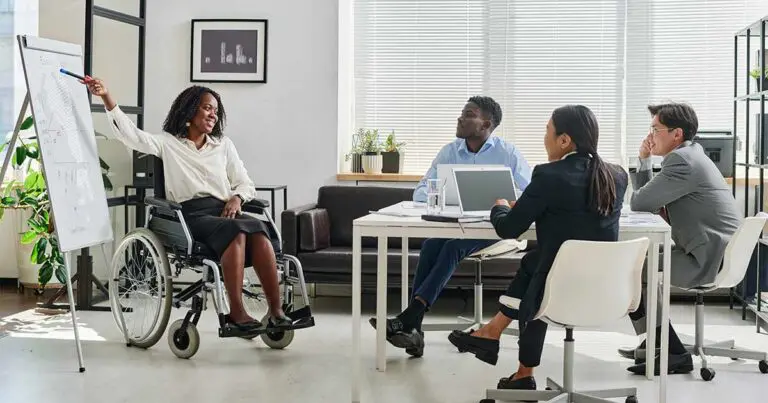 A diverse group of professionals is engaged in a business meeting in a modern office setting. A woman in a wheelchair is presenting information on a whiteboard to three colleagues who are seated at a table with laptops and notebooks. The room is well-lit with natural light coming through large windows, and there are plants and modern office furniture visible in the background.