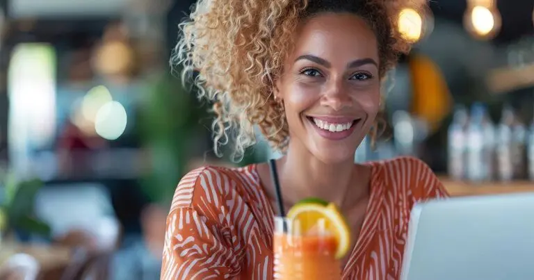 A young woman with curly hair smiles at the camera while sitting in a cafe. She is wearing a vibrant orange and white patterned shirt and working on a laptop. In front of her, there is a colorful drink garnished with a slice of orange. The background features a cozy cafe setting with warm lighting and blurred patrons.