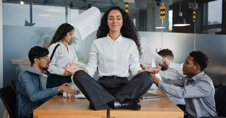 A woman is seated cross-legged on top of a desk in a busy office, practicing meditation with her eyes closed and a calm expression. Around her, coworkers appear stressed, with papers flying and gestures indicating a hectic environment, highlighting the contrast between her serene state and the surrounding chaos to symbolize the importance of employee wellbeing programs