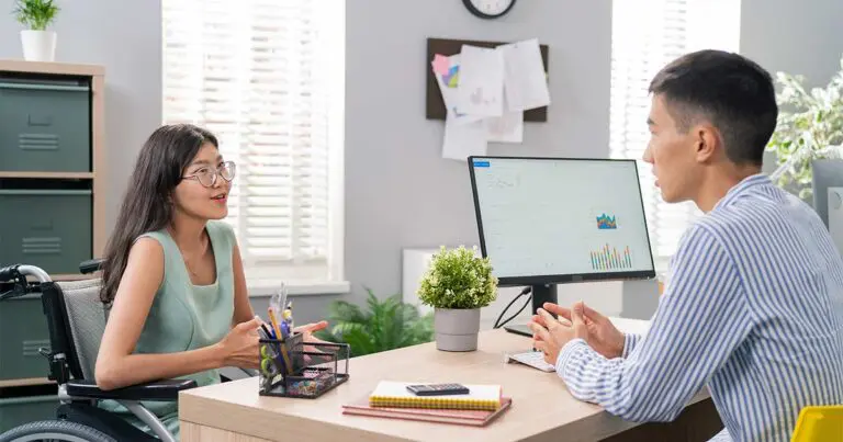 A woman in a wheelchair is seated at a desk during a job interview. She appears engaged and confident as she talks to the interviewer, who is sitting across from her. The setting is a modern office with a computer monitor displaying graphs and charts. This scene exemplifies a professional scenario where candidates might be asked, "How do you handle stress and pressure?"