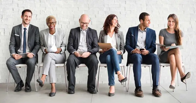 six diverse professionals seated in a row against a white brick wall, waiting for an interview or meeting. Each person is dressed in business attire, and they appear to be engaged in various activities such as reviewing documents, using a tablet, or conversing with one another. The individuals represent a range of ages, genders, and ethnicities, reflecting a diverse and inclusive workplace setting to represent building a talent pipeline