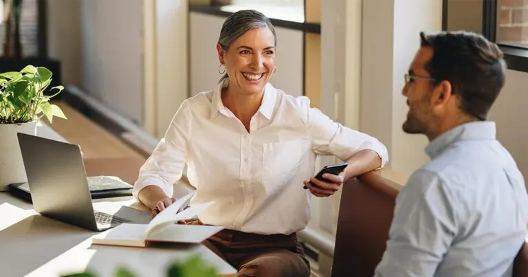 A professional woman smiling and conducting a mock interview with a man in an office setting, using mock interview questions to prepare him for a job interview.