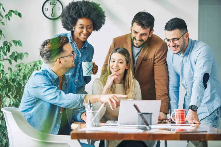 A diverse group of five colleagues is gathered around a desk in an office, smiling and collaborating while looking at a laptop. The group includes men and women, all casually dressed, with one person pointing at the laptop screen while the others engage attentively. A clock and potted plant are visible in the background, adding to the relaxed and productive atmosphere