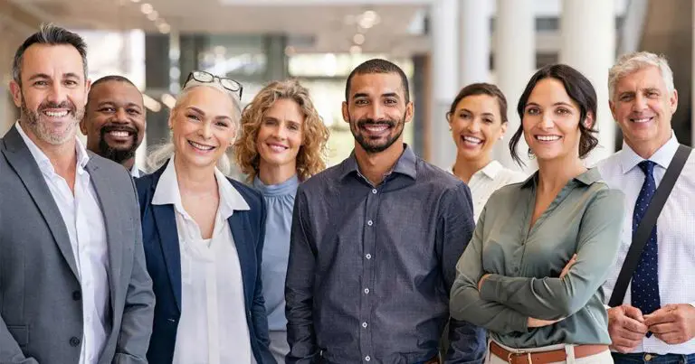 A diverse group of professionals smiling confidently while standing together in a bright, modern office environment. The group includes men and women of various ages and ethnicities, reflecting teamwork and inclusivity in a professional setting.