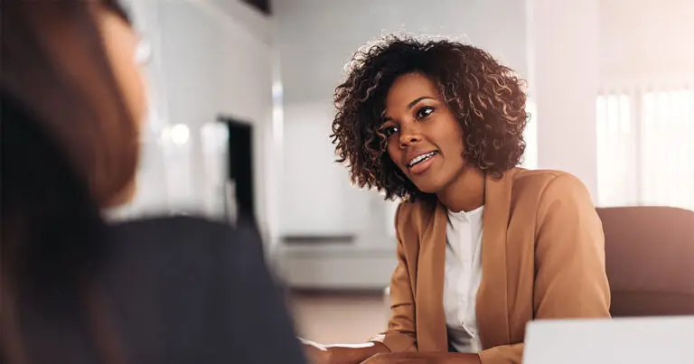 The image shows a professional setting where a woman with curly hair is attentively listening to another person. The woman is wearing a light brown blazer over a white top, and her body language suggests she is engaged in the conversation, possibly during an interview or a meeting. The room is softly lit, and the focus is on the woman's thoughtful expression to symbolize how to answer How do you handle criticism in an interview