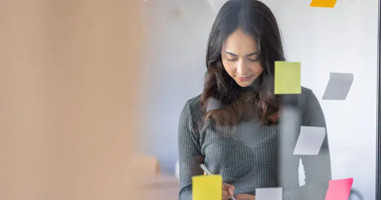 Young professional woman working in an office, writing notes on a glass wall covered in colorful sticky notes, deep in thought as she organizes her tasks to represent how to answer how do you prioritize your work in an interview