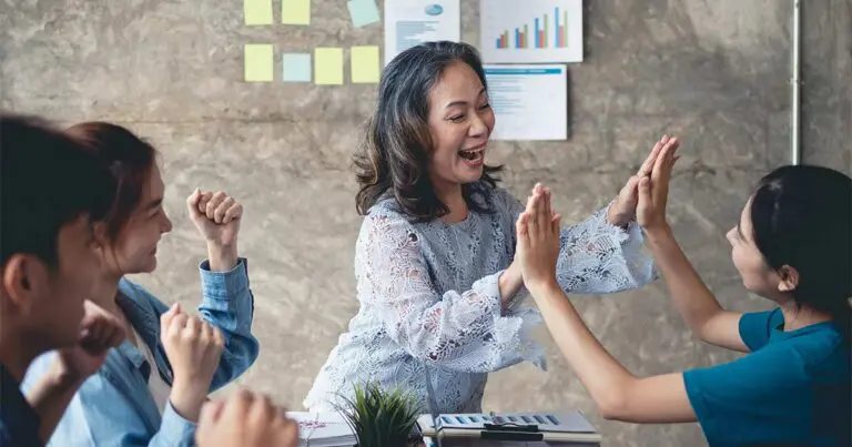 A group of diverse colleagues, including an older woman and younger individuals, happily high-five each other in a workplace setting. They are seated around a table with charts and sticky notes on the wall in the background, suggesting a collaborative or celebratory moment during a team meeting.