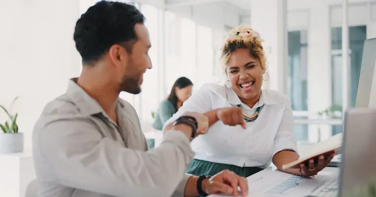 A cheerful office setting where two colleagues, one holding a notebook, are laughing and engaging in a lively discussion, fostering a positive and collaborative work environment to symbolize how to answer what motivates you in an interview