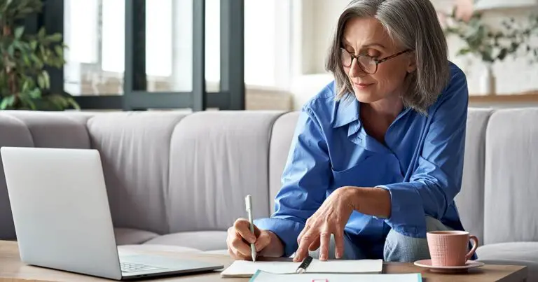 A mature woman wearing glasses sits on a sofa, focused on taking notes while working on a laptop. A cup of coffee is placed beside her on the table. This image can represent someone preparing for a remote job interview, making it ideal for content on how to answer "why do you want to work remote" in an interview.