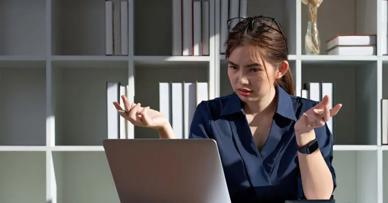 The image shows a woman sitting in front of a laptop, appearing confused or frustrated. She has her hands slightly raised in a questioning gesture, as if perplexed by what she sees on the screen. Her expression is serious, and she is wearing a dark blue blouse, with glasses resting on her head. Shelves with books are visible in the background, adding a professional or office-like setting.