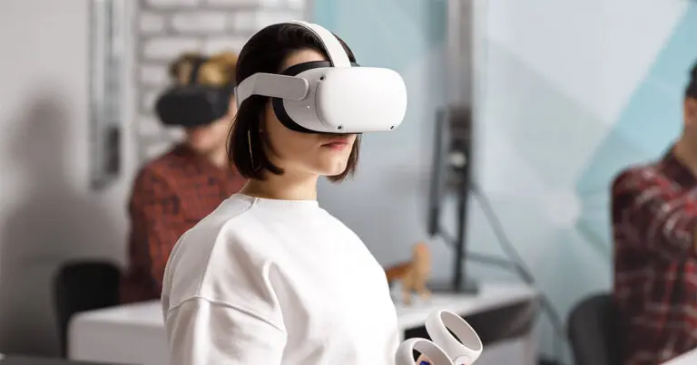 The image shows a person wearing a virtual reality (VR) headset in a modern office or training environment. The individual, dressed in a white top, is holding VR controllers in their hand, appearing focused on an immersive experience. In the background, another person, also wearing a VR headset, is blurred but appears to be engaged in a similar activity. The setting suggests the use of VR technology for purposes such as employee training or development in a professional context.