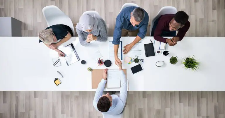 A diverse group of interviewers seated around a white conference table, reviewing documents while one interviewer shakes hands with a candidate, signaling the conclusion of a successful interview to symbolize the growing popularity of gig-work and how staffing agencies can help