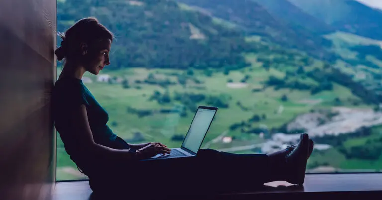 Image of a woman sitting in front of a beautiful natural backdrop of rolling green hills and mountains with a laptop on her lap to symbolize the rise in digital nomadism.