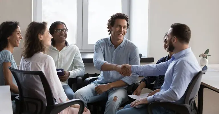 A diverse group of professionals sitting around in a semi-circle in office chairs in an office setting. Two of the professionals are shaking hands as to congratulate each other while their colleagues smile in appreciation all to represent building effective employee referral programs.