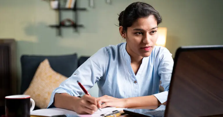 A professional woman sitting at a desk looking at a computer screen and taking notes in a notebook to symbolize how to answer How do you stay current in your field in an interview.