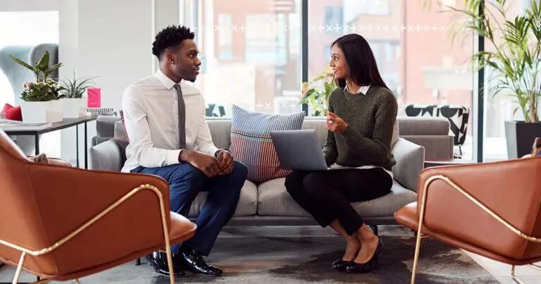 A man and woman are seated on a modern office couch, engaged in a conversation. The man is wearing a white shirt and tie, while the woman, holding a laptop, is dressed in a green sweater and black pants. The office space has comfortable chairs, plants, and natural light coming through large windows, creating a professional yet relaxed atmosphere