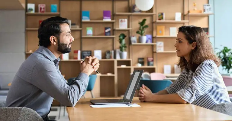 The image shows two people sitting across from each other at a wooden desk in a modern, open office or library-like setting. The person on the left, a man with a beard, is attentively listening with his hands clasped together in front of him. The woman on the right, dressed in a striped shirt, sits with her hands resting on the table, speaking and making eye contact. Between them, a laptop and some papers are placed on the table. The background includes bookshelves filled with books and small plants, creating a professional yet relaxed atmosphere