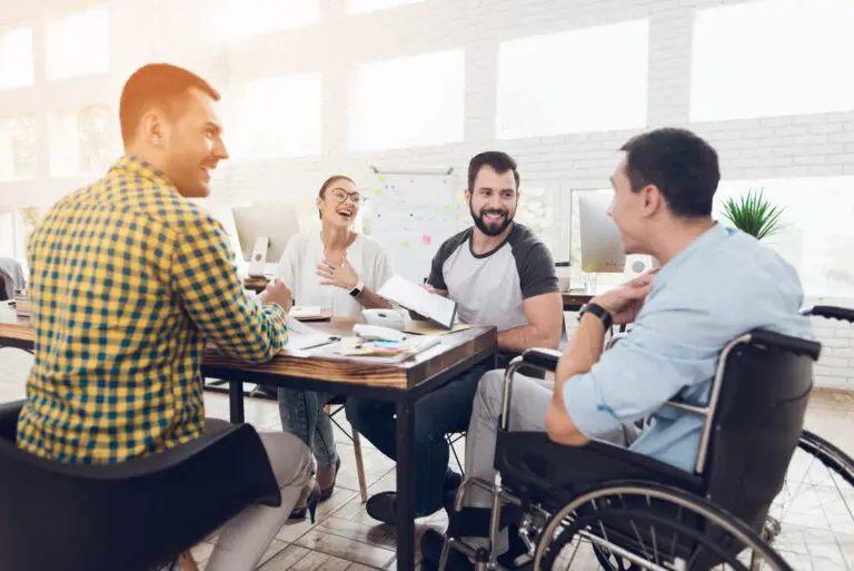 Alt text: A diverse group of colleagues is gathered around a table in a bright, modern office space, engaged in a lively discussion. One person in a wheelchair smiles and speaks with another individual across the table. The group appears relaxed and happy, with papers, notebooks, and coffee cups spread out on the table, indicating a collaborative work environment. The setting suggests inclusivity and teamwork, with a whiteboard in the background featuring colorful sticky notes to symbolize creativity in tech teams.