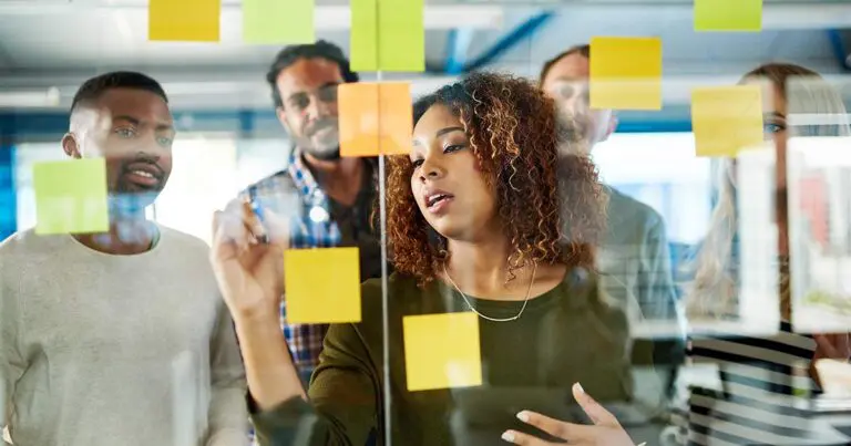 A diverse group of professionals standing in front of a glass wall covered in multi-colored post-it notes while discussing their strategy to symbolize the difference between marketing vs sales enablement