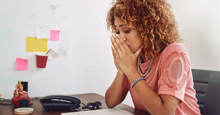 A professional woman in a pink blouse sitting at her desk with her hands tented around her face with a look of distress to symbolize the feeling of "my team was laid off. now what?"