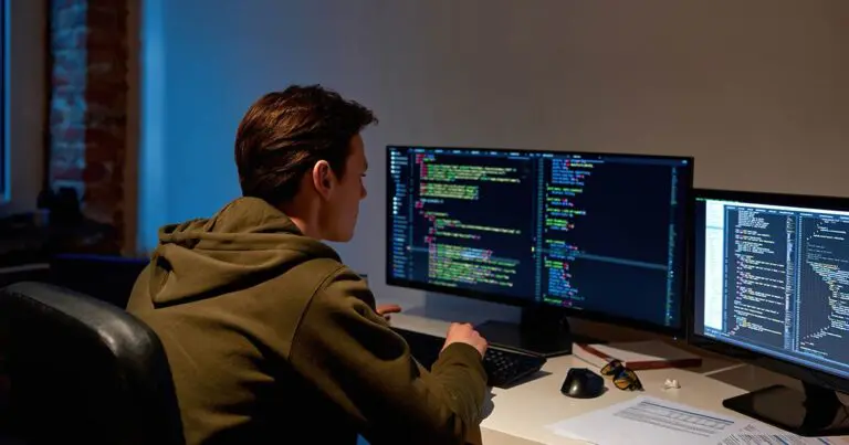 A man with short hair wearing a brown hooded sweatshirt sitting in a dimly lit room at a desk with two computer monitors with what appears to be lines of code on the screens to represent remote work cybersecurity challenges