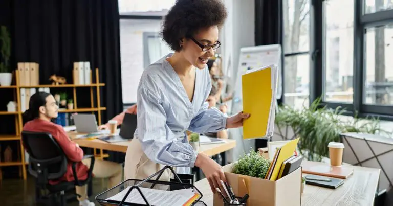 A professional woman in a blue shirt happily packing up her things from her desk at work to symbolize making a career transition to move to a new industry