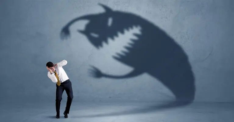 A man in business attire cowers in fear as a large, monstrous shadow with sharp teeth and claws looms over him, symbolizing the intimidating and daunting aspects of hiring someone new.