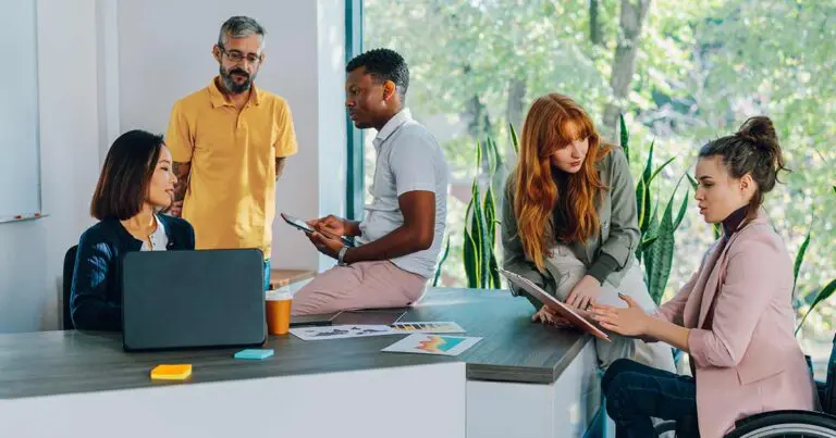 A diverse group of professionals working in an office setting with a large picture window sitting around an office table working together to symbolize an agile workforce