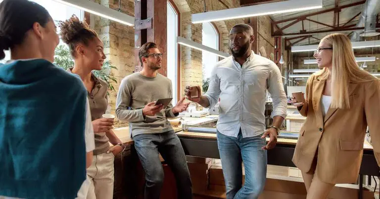 A diverse group of professionals in an office space bathed in natural sunlight all standing around in a semi-circle in a casual fashion to represent answering what's your leadership style in an interview