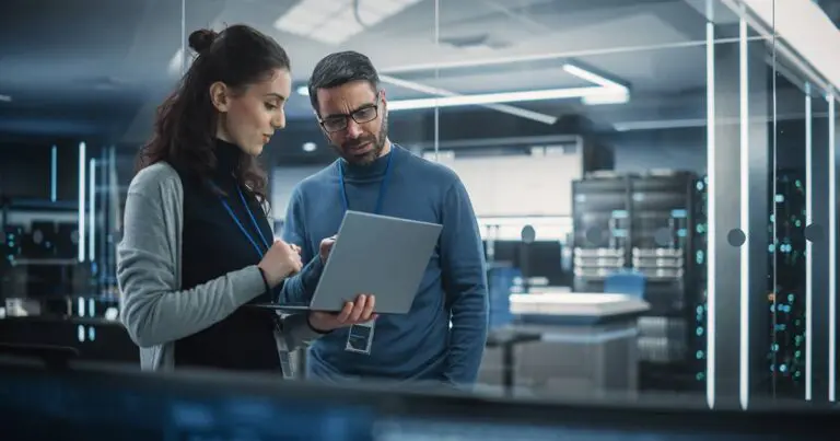 A woman and a man stand in a dimly lit office looking at a laptop the woman is holding.