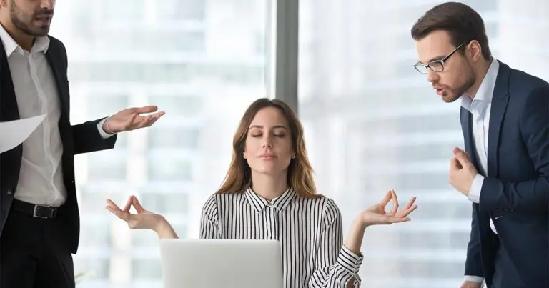 A woman sits at her desk with her eyes closed and hands in a meditative pose, remaining calm while two male colleagues on either side argue and gesture around her in a busy office environment.