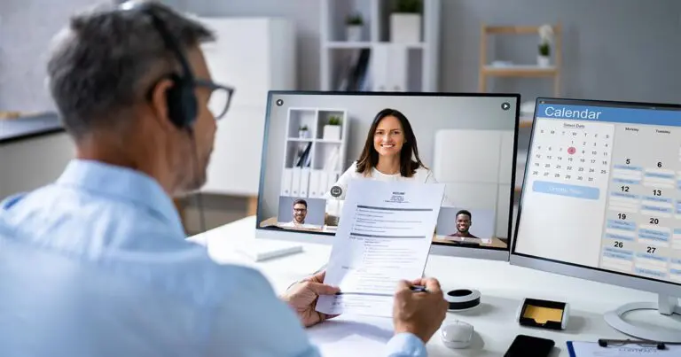 A man in a professional setting conducts a virtual job interview. He holds a printed resume while looking at a video conference screen displaying a female candidate in the center and two other small video feeds of participants at the bottom. A second monitor shows a calendar with scheduled tasks to symbolize how to answer "What Are Your 30-60-90 Day Plans?" in an Interview