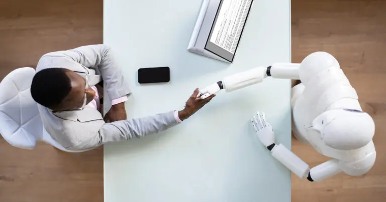 Bird's-eye view of a professional in a business suit shaking hands with a humanoid robot across a desk, symbolizing LinkedIn's AI hiring assistant as a game-changer for recruiters and hiring managers. A tablet and smartphone are visible on the desk, enhancing the modern, tech-forward atmosphere.