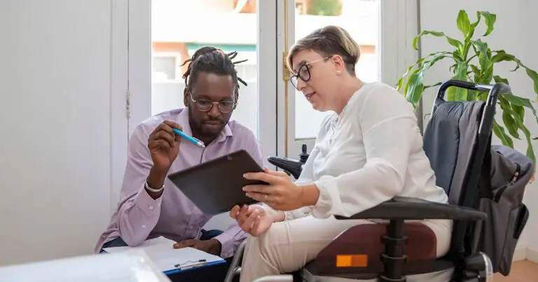 A man and a woman are sitting together in an office space discussing something on a tablet. The man has dreadlocks, glasses, and is wearing a light purple shirt while holding a pen. The woman, sitting in a wheelchair, is wearing a white blouse, glasses, and is holding the tablet as they engage in conversation. A green potted plant is visible in the background, and the room is well-lit with natural light streaming through a window.