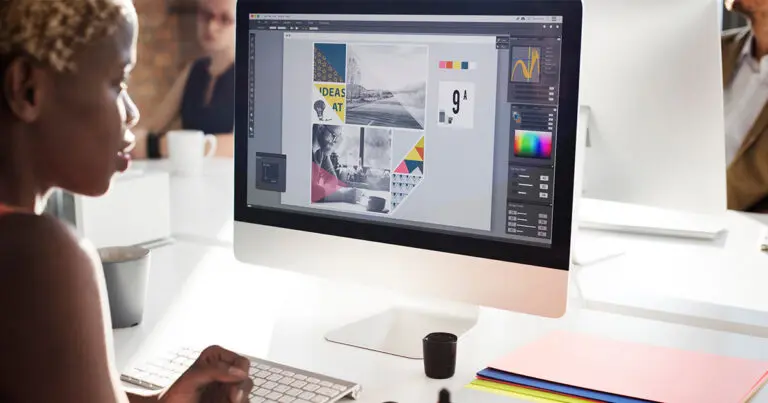 Person working on graphic design software on a desktop computer in a modern office setting, with a colorful design project displayed on the screen. A colleague is visible in the background.