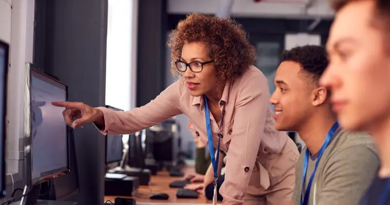 A professional woman wearing glasses and a lanyard leans over to point at a computer screen while guiding two young adults seated at computers in a modern classroom or training environment.