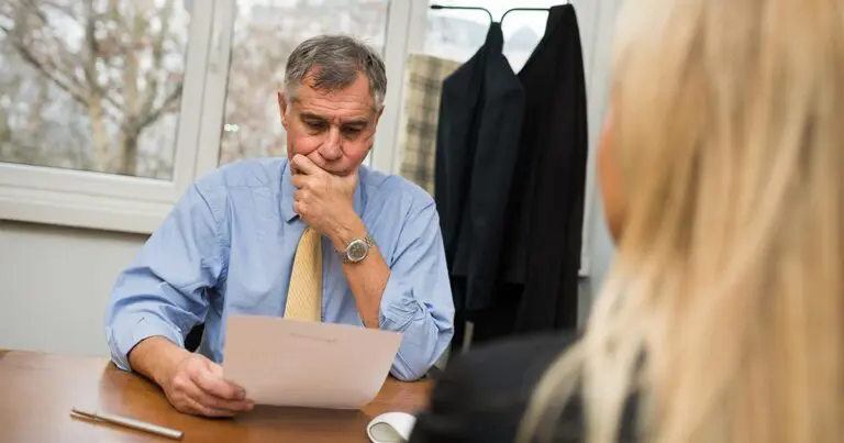 A middle-aged man with short gray hair, wearing a light blue dress shirt and a yellow tie, sits at a desk reviewing a document with a thoughtful expression. His hand rests on his chin as he looks at the paper. A woman with long blonde hair, seen from behind, sits across from him. The setting appears to be a professional office with a coat rack holding jackets in the background and large windows showing trees outside.