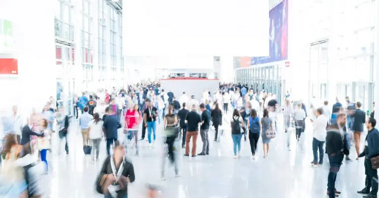 The image depicts a busy tech expo like CES 2025 with a large crowd of people walking through a spacious, brightly lit exhibition hall. Attendees are dressed in a mix of business and casual attire, some carrying bags or wearing lanyards. The background features large glass windows allowing natural light to flood the space, with various booths and banners visible along the sides of the hall. The motion blur effect gives the impression of movement and a bustling atmosphere.